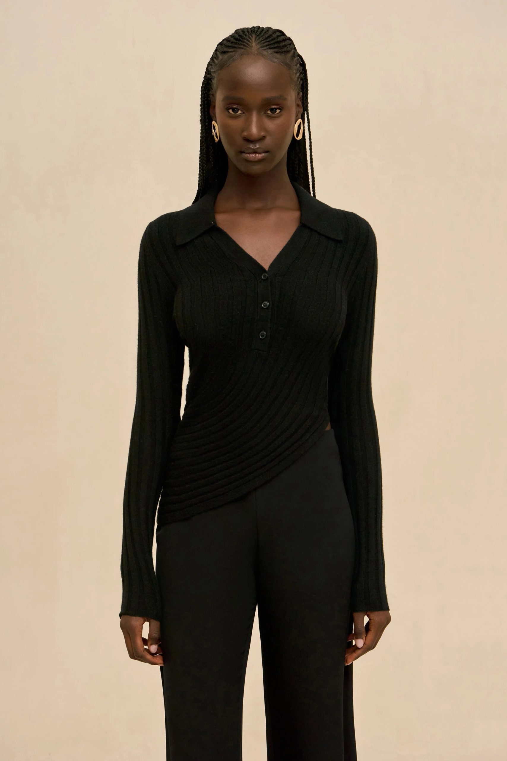 Image 410 - New Trend Dark-skinned woman in a black ribbed knit top with a V-neck and buttons, wearing gold hoop earrings, standing against a beige background.