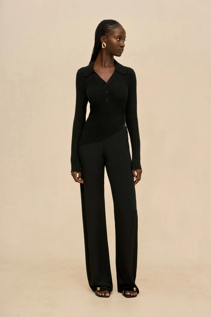Elegant Black woman in a black ribbed knit blouse and wide-leg trousers stands against a beige background.