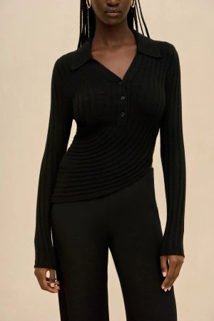 Person wearing a black ribbed knit top with buttons and a V-neck, paired with black pants, standing against a beige background, visible braided hair and hoop earrings.