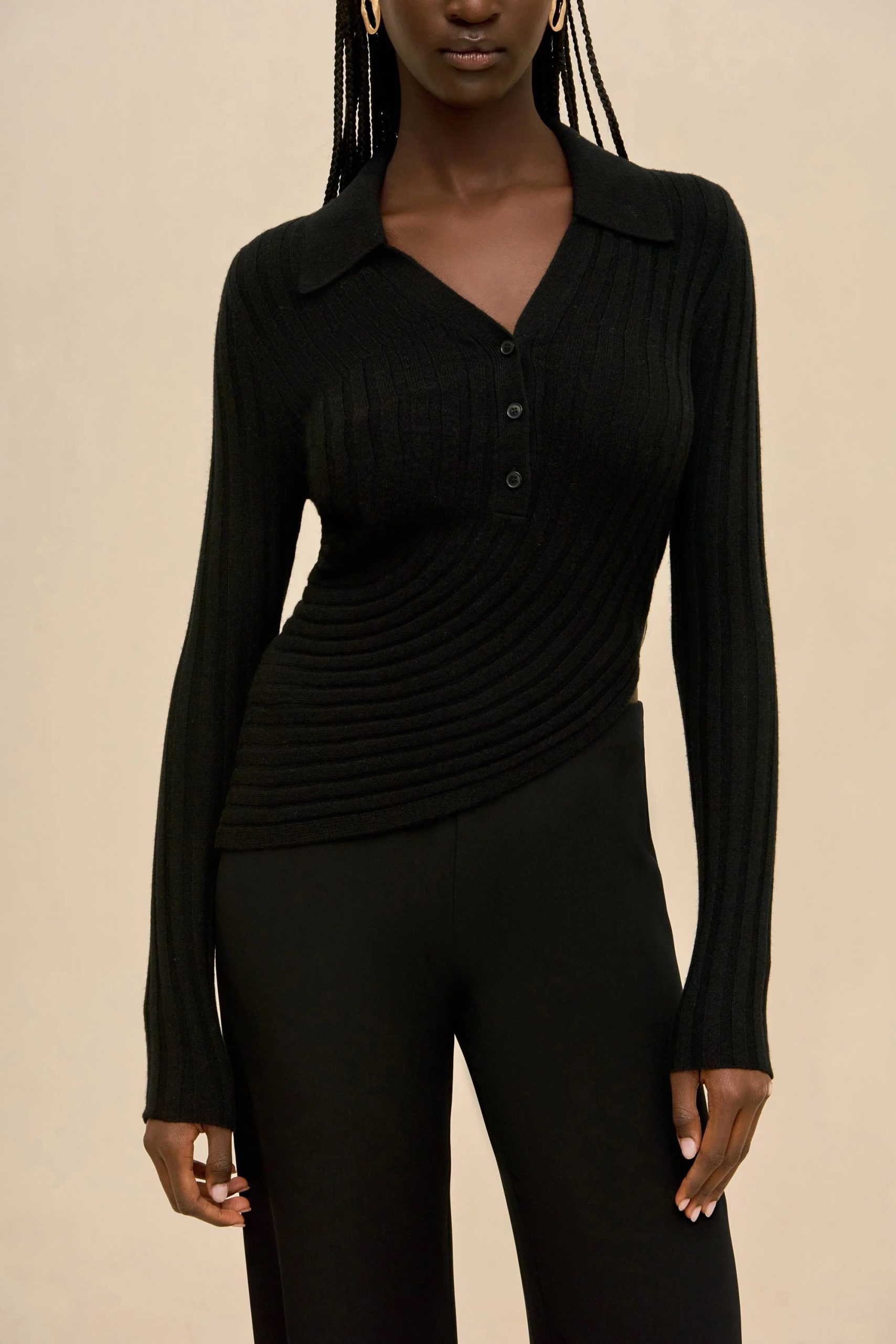 Image 412 - New Trend Person wearing a black ribbed knit top with buttons and a V-neck, paired with black pants, standing against a beige background, visible braided hair and hoop earrings.