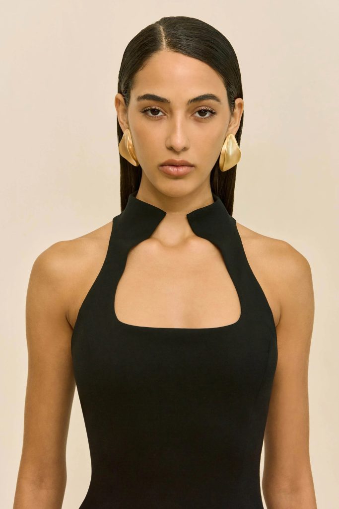 Portrait of a woman with straight dark hair, wearing a black halter-neck dress and oversized gold earrings against a neutral beige background.