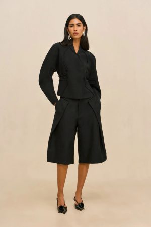 Woman modeling a black structured blazer and wide-leg shorts, hands in pockets, neutral backdrop.