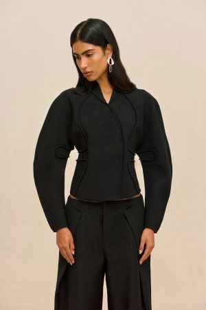 Fashion-oriented portrait of a woman in a black, structured jacket with decorative seam details, wearing large white geometric earrings against a beige backdrop, looking down slightly to the left.