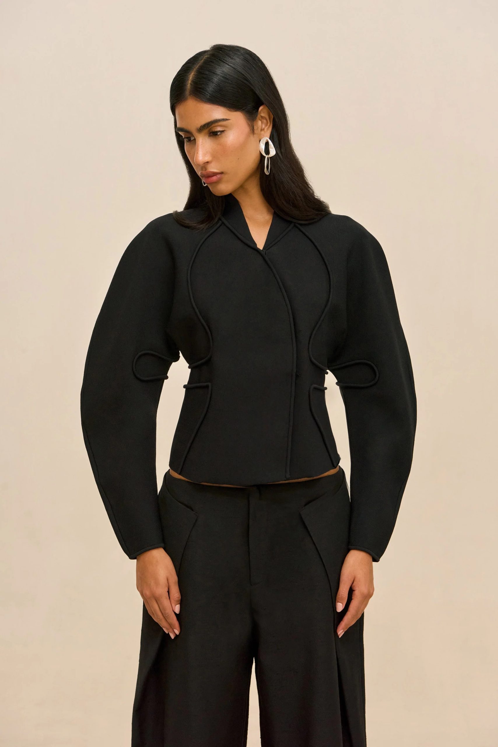 Image 421 - New Trend Fashion-oriented portrait of a woman in a black, structured jacket with decorative seam details, wearing large white geometric earrings against a beige backdrop, looking down slightly to the left.