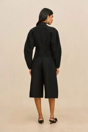 Back view of a woman in a black tailored outfit with sculptural back seam details, wearing black sandals, neutral beige background.