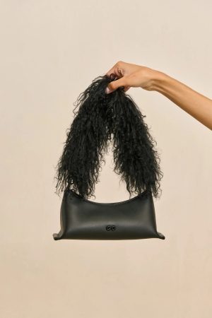 Hand lifting a bunch of long, curly black hair above a black leather bag against a beige background.