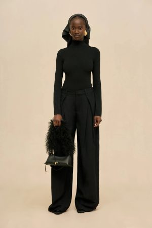 Full-length portrait of a woman in a black turtleneck and wide-leg pants, wearing a black headscarf and gold earrings, holding a black feathered handbag with a small gold logo.
