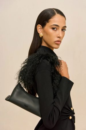 Woman in a black outfit with a large black shoulder bag, gold circular earring, and neutral beige background (fashion portrait).