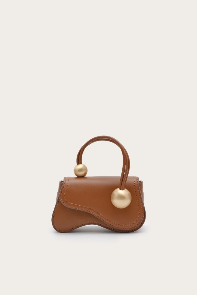 Brown leather handbag with curved top, double handles, and large gold sphere accents on the handles against a light background.