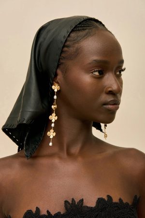Close-up of a woman with dark skin wearing a black headscarf and gold floral earrings, facing right against a beige background, profile view.