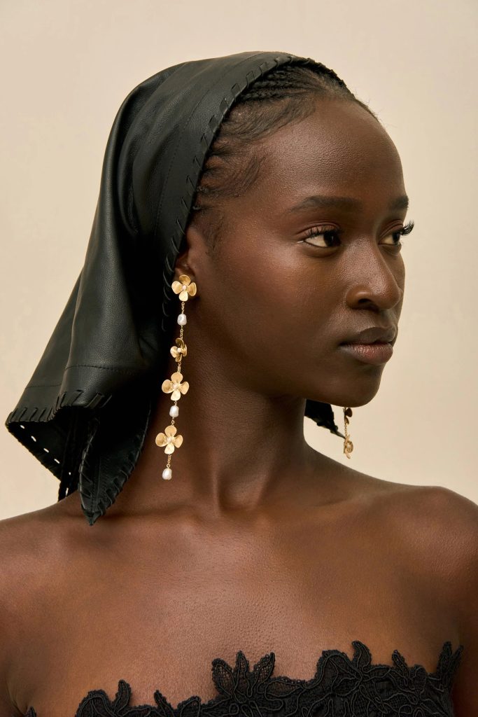 Close-up of a woman with dark skin wearing a black headscarf and gold floral earrings, facing right against a beige background, profile view.
