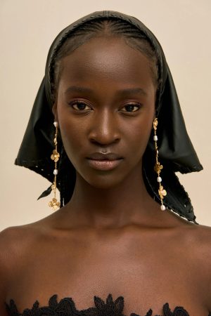 Close-up portrait of a young woman wearing a black headscarf and gold earrings, facing the camera against a beige background.