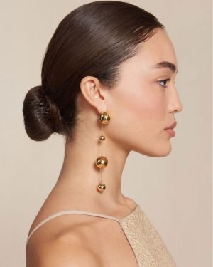 Side profile of a woman with a sleek bun wearing a long gold bead drop earring in beige tones.