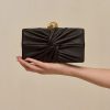 Black leather clutch with a twisted knot front detail, held in a hand against a beige background.