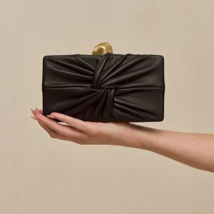 Black leather clutch with a twisted knot front detail, held in a hand against a beige background.