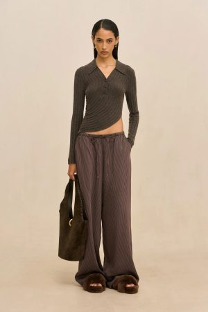 Full-body view of a woman wearing a dark cropped ribbed top, striped wide-leg pants, brown fuzzy slippers, and carrying a brown bag against a beige backdrop.