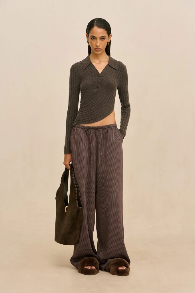 Full-body view of a woman wearing a dark cropped ribbed top, striped wide-leg pants, brown fuzzy slippers, and carrying a brown bag against a beige backdrop.