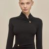 Model with slicked-back blonde hair wearing a black top with gold buttons, standing against a neutral beige backdrop.