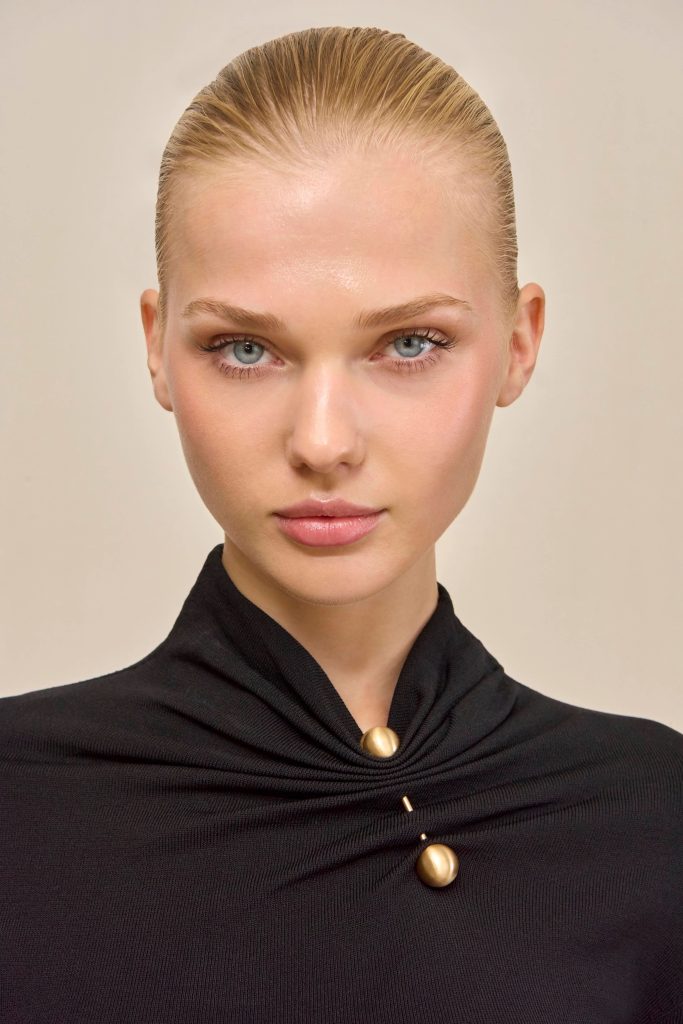 Close-up portrait of a woman with slicked-back blonde hair, blue eyes, and natural makeup wearing a black garment with a gold bead necklace cap
