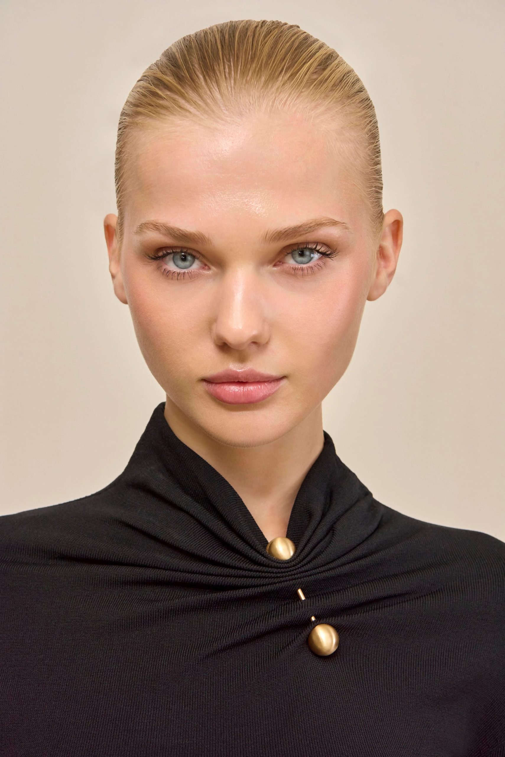 Image 455 - New Trend Close-up portrait of a woman with slicked-back blonde hair, blue eyes, and natural makeup wearing a black garment with a gold bead necklace cap