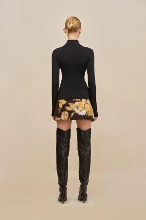Back view of a person wearing a fitted black long-sleeve top, a floral mini skirt, and tall black boots against a beige backdrop, with hair styled in a bun.