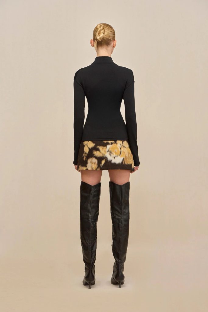 Back view of a person wearing a fitted black long-sleeve top, a floral mini skirt, and tall black boots against a beige backdrop, with hair styled in a bun.