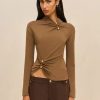 Model wearing a taupe long-sleeve top with twisted front detail and gold buttons, paired with dark brown pants against a neutral backdrop.
