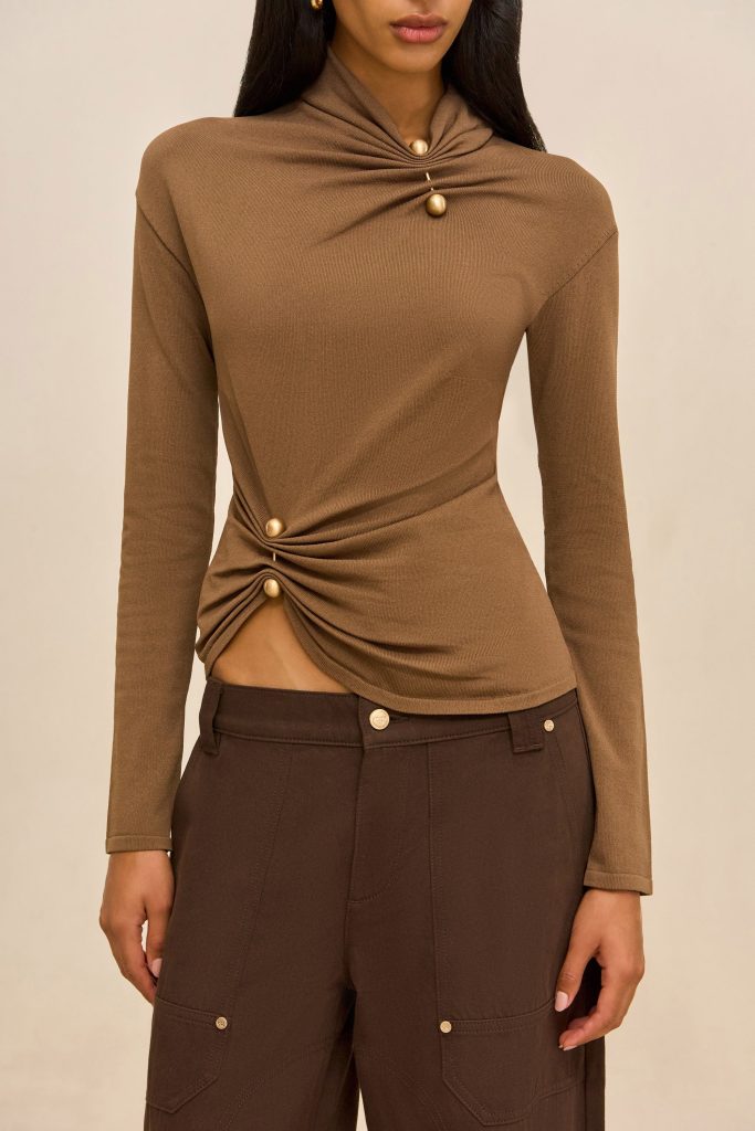 Woman wearing a brown long-sleeve knit top with a twisted knot detail at the waist and gold buttons, paired with dark brown pants.