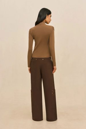 Back view of a woman wearing a tan turtleneck and dark brown wide-leg pants with pockets, standing against a beige background.