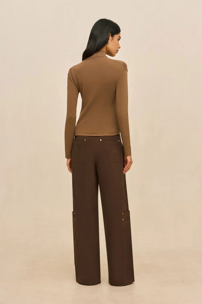 Back view of a woman wearing a tan turtleneck and dark brown wide-leg pants with pockets, standing against a beige background.
