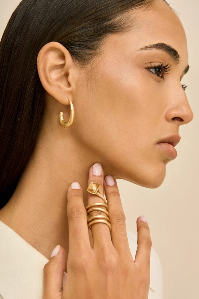Close-up side profile of a woman wearing gold jewelry: hoop earring and sculptural rings, hand resting by jawline.