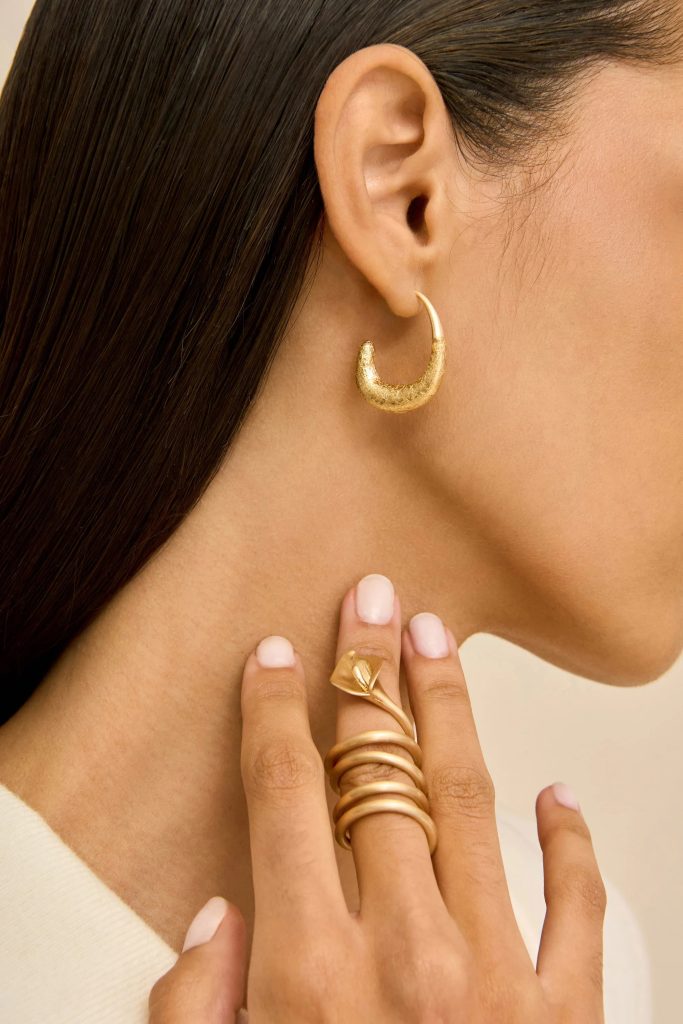 Close-up of a woman's ear wearing a textured gold crescent hoop earring, with multiple gold rings and pale-pink nails nearby on the hand.