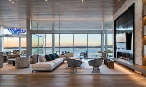 Open-concept living room with a large sectional sofa and woven chairs, facing floor-to-ceiling windows with an ocean view at sunset.