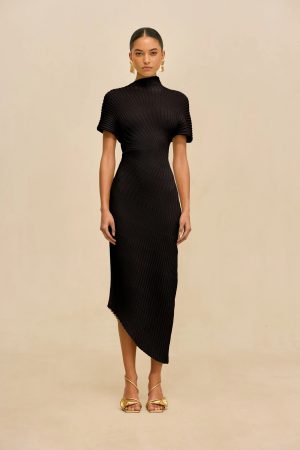 Full-body shot of a woman in a black ribbed knit dress with a high neck, short sleeves, and an asymmetric hem, wearing gold sandals and statement earrings against a beige background.