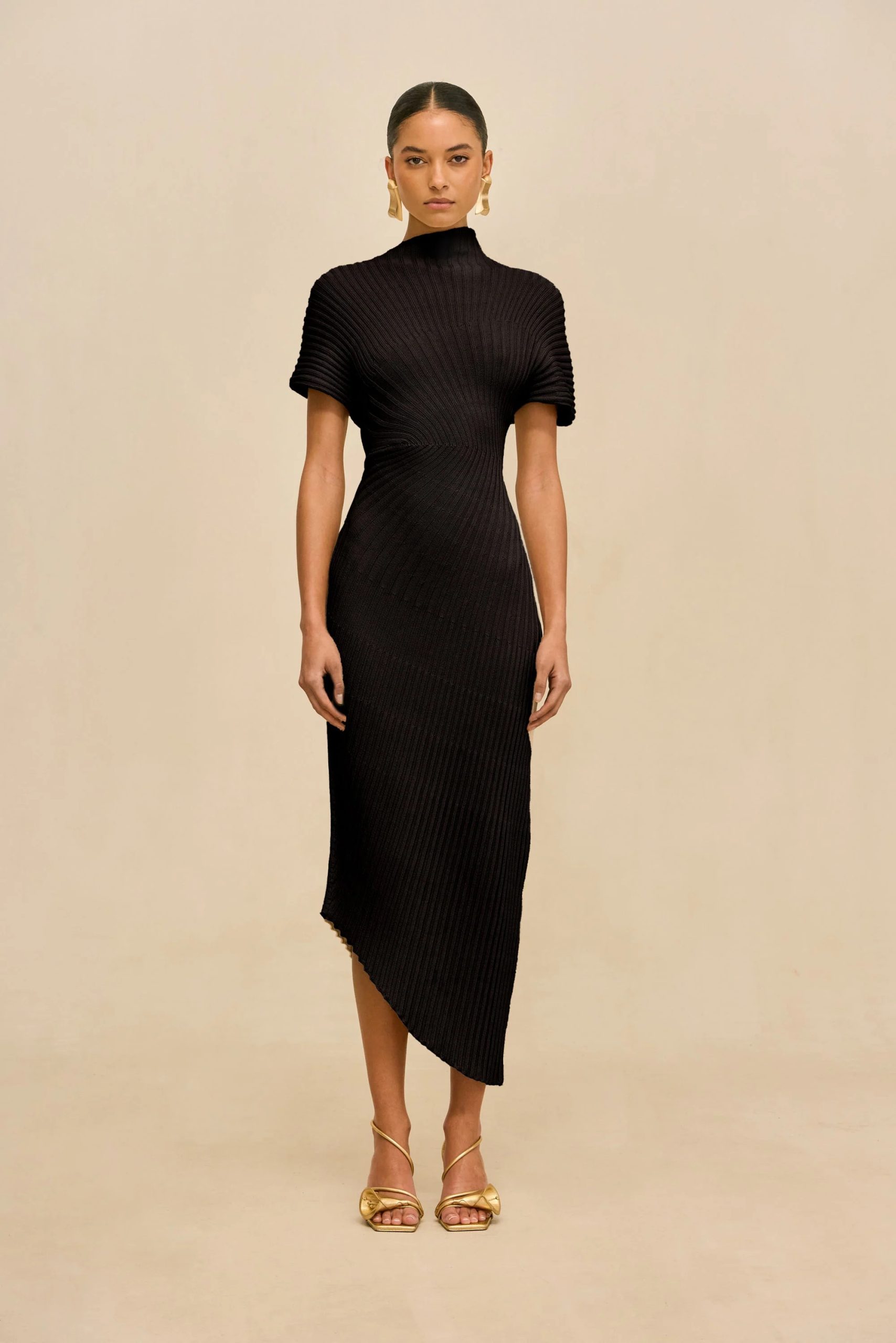 Image 474 - New Trend Full-body shot of a woman in a black ribbed knit dress with a high neck, short sleeves, and an asymmetric hem, wearing gold sandals and statement earrings against a beige background.