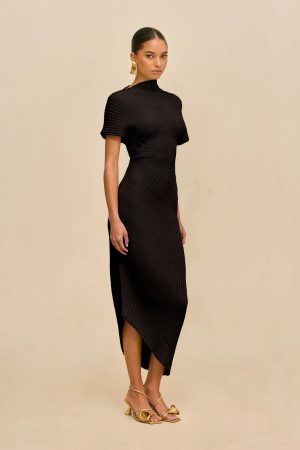 Woman wearing a black ribbed dress with a high neck and asymmetrical hem, standing against a beige backdrop, gold earrings and sandals visible.