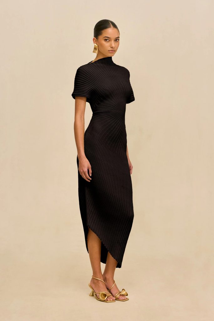 Woman wearing a black ribbed dress with a high neck and asymmetrical hem, standing against a beige backdrop, gold earrings and sandals visible.