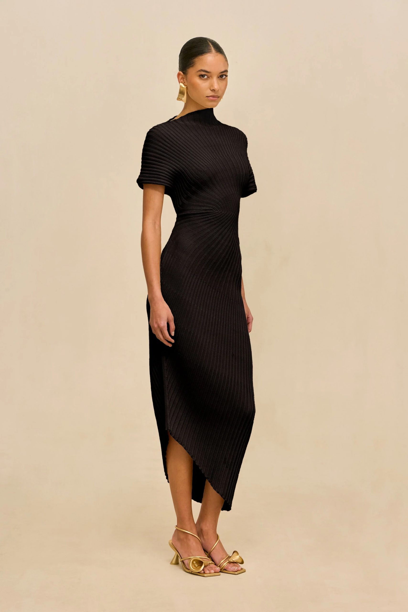 Image 476 - New Trend Woman wearing a black ribbed dress with a high neck and asymmetrical hem, standing against a beige backdrop, gold earrings and sandals visible.
