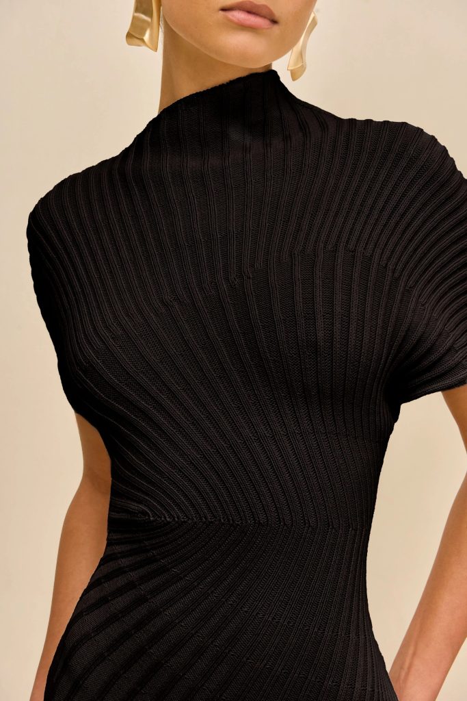 Close-up of a person in a black ribbed knit top with a high neck against a beige background.