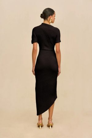 Back view of a woman in a black ribbed knit midi dress, standing against a beige background, with her hair in a bun and gold heeled sandals.
