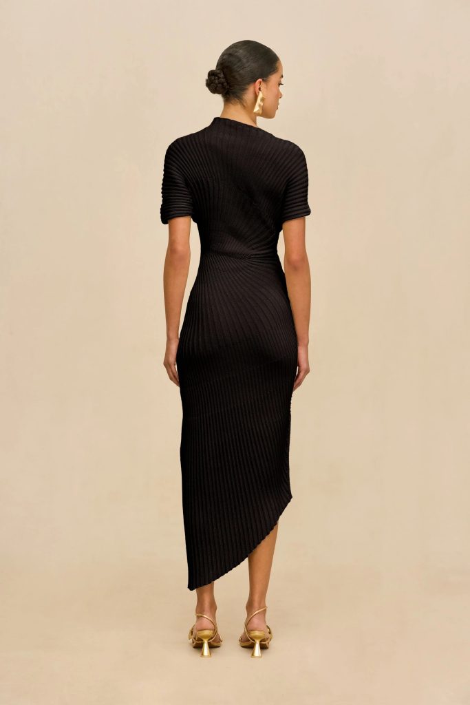 Back view of a woman in a black ribbed knit midi dress, standing against a beige background, with her hair in a bun and gold heeled sandals.