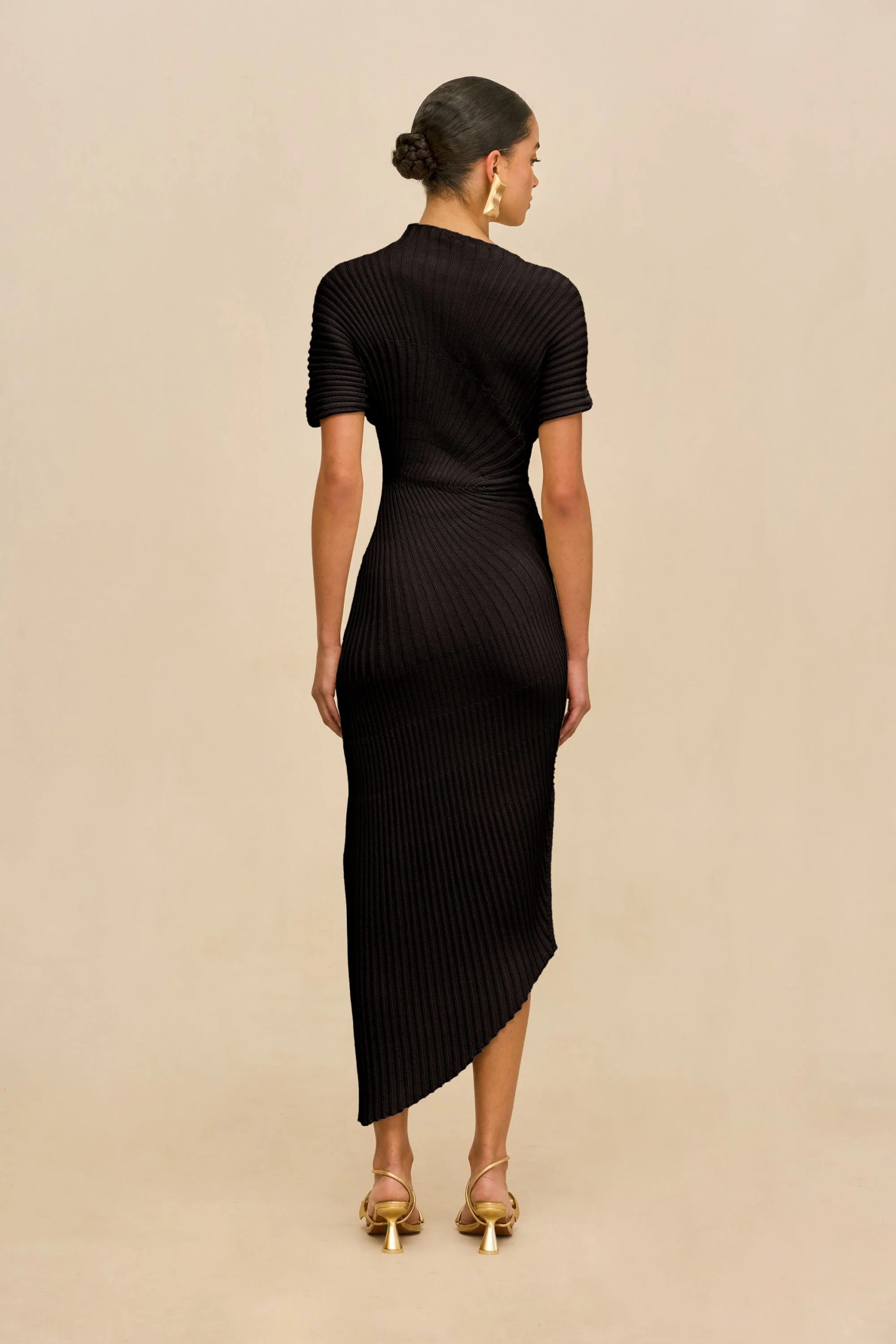 Image 478 - New Trend Back view of a woman in a black ribbed knit midi dress, standing against a beige background, with her hair in a bun and gold heeled sandals.