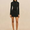 Image 479 - New Trend Model wearing a black long-sleeve mini dress with twisted front and gold button details, standing against a neutral beige backdrop with strappy sandals.