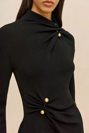 Close-up of a person wearing a black knit top with a folded, draped neckline and gold round buttons at the chest area against a beige background.