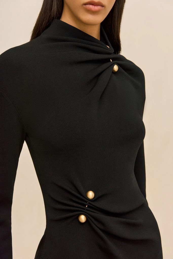Close-up of a person wearing a black knit top with a folded, draped neckline and gold round buttons at the chest area against a beige background.