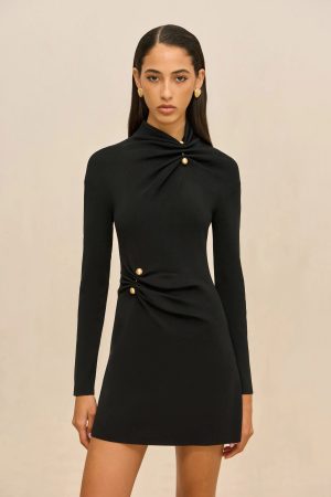 Woman wearing a black, long-sleeve dress with gold button accents on the neckline and waist, standing against a neutral background.
