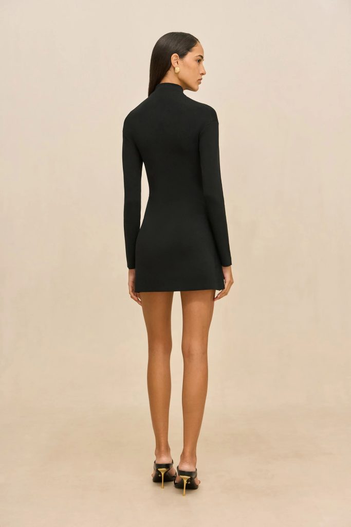 Back view of a woman wearing a fitted black long-sleeve mini dress, standing against a beige background.