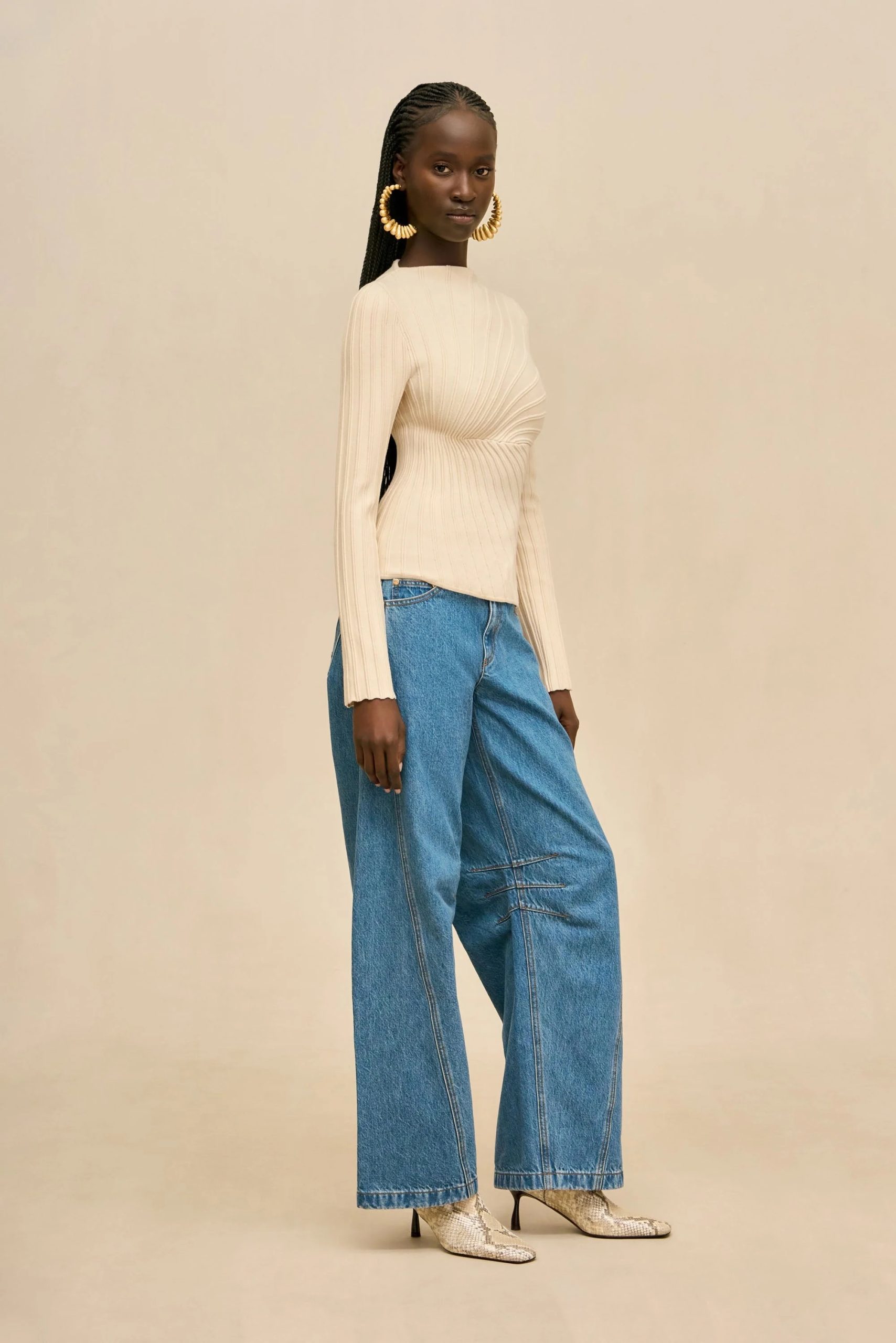 Image 494 - New Trend Model wearing a cream ribbed knit top with wide-leg blue jeans, snakeskin heeled boots, and large hoop earrings, posed against a beige backdrop.