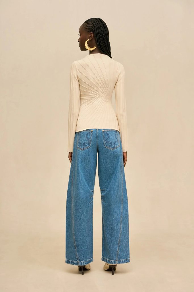 Back view of a woman wearing a beige ribbed long-sleeve top tucked into high-waisted wide-leg blue jeans, with braided hair and large circular gold earrings.