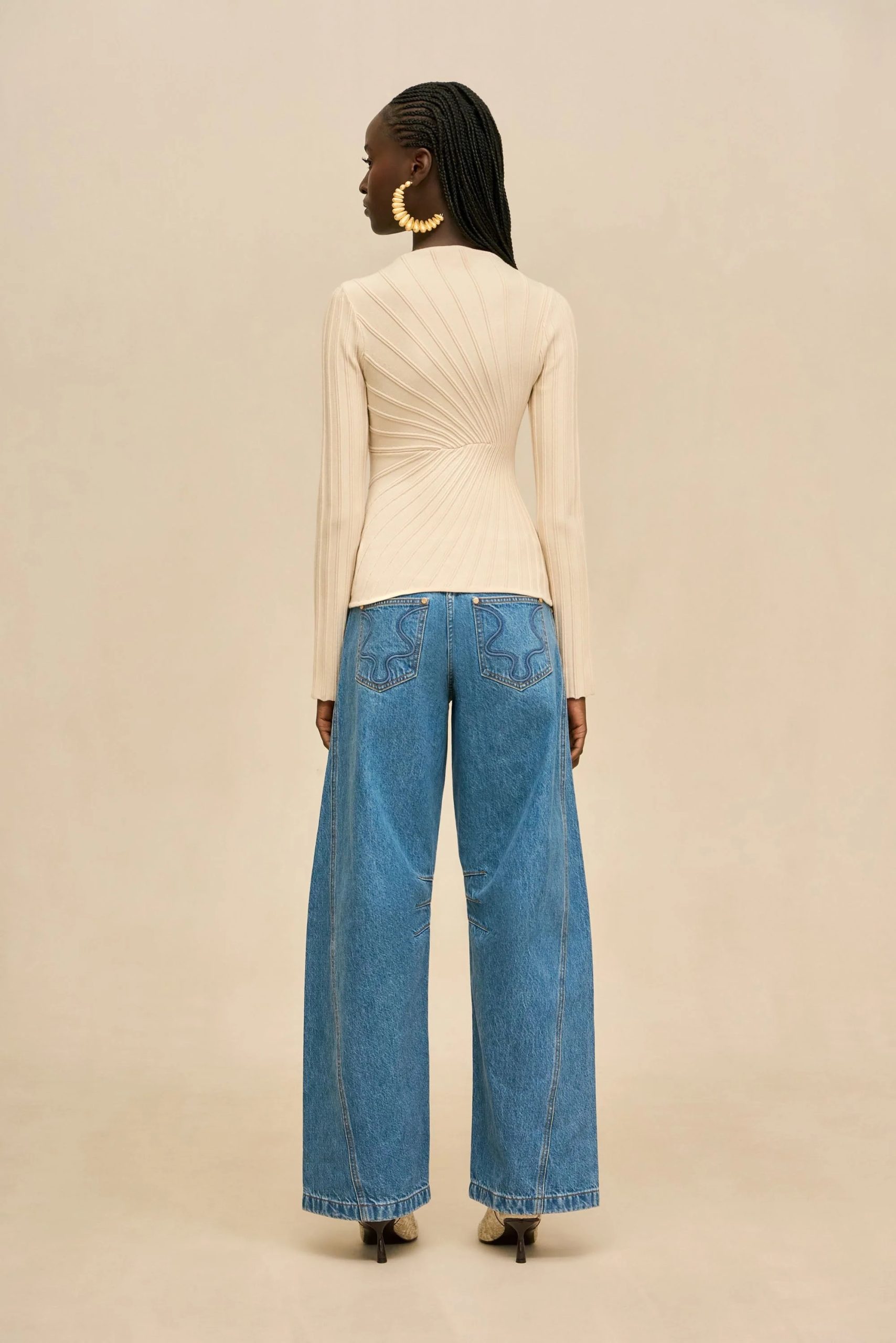 Image 495 - New Trend Back view of a woman wearing a beige ribbed long-sleeve top tucked into high-waisted wide-leg blue jeans, with braided hair and large circular gold earrings.